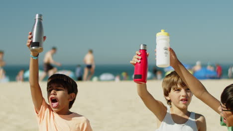 Fröhliche-Sportler-Sitzen-Am-Strand-Und-Heben-Wasserflaschen-Hoch